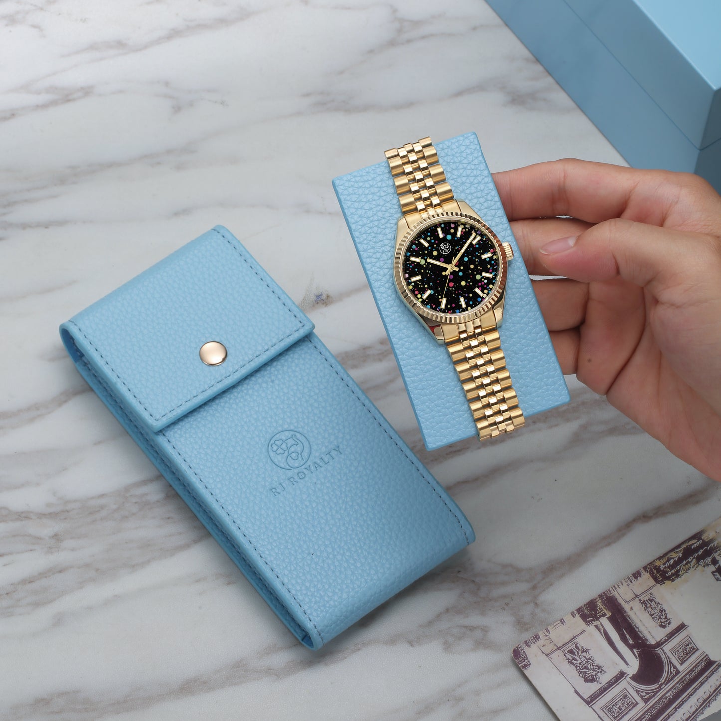 A luxury leather watch case in blue, shown holding a gold-banded watch. The case is presented with a visible RJ Royalty brand logo on the bottom right.