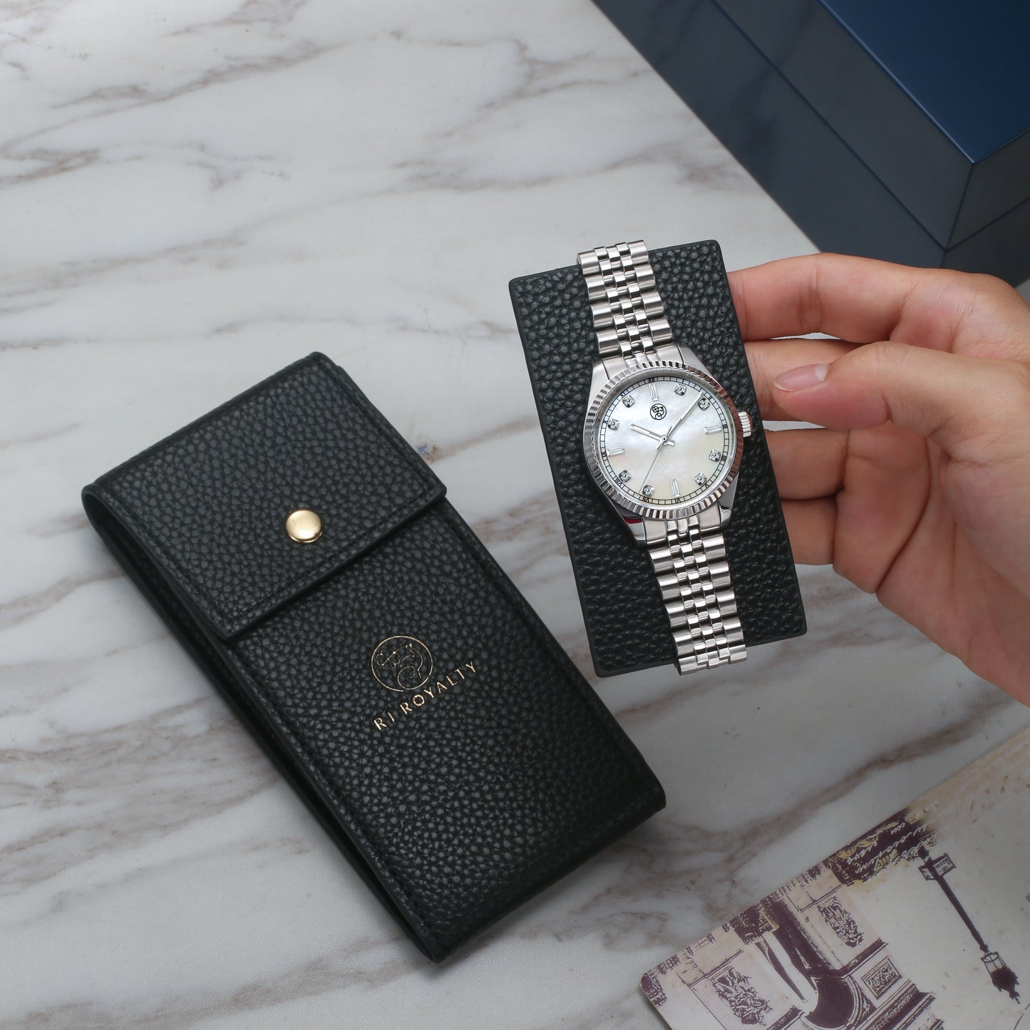 A black luxury watch case and a hand holding a silver watch with a mother of pearl dial with baguette diamond markers.